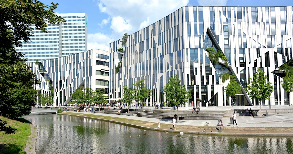 Grün trifft Architektur: Moderne Glasfassade, im Vordergrund ein ruhiger Fluss, in dem sich eine gepflegte Grünanlage spiegelt, links und rechts Teile weiterer Gebäude sichtbar